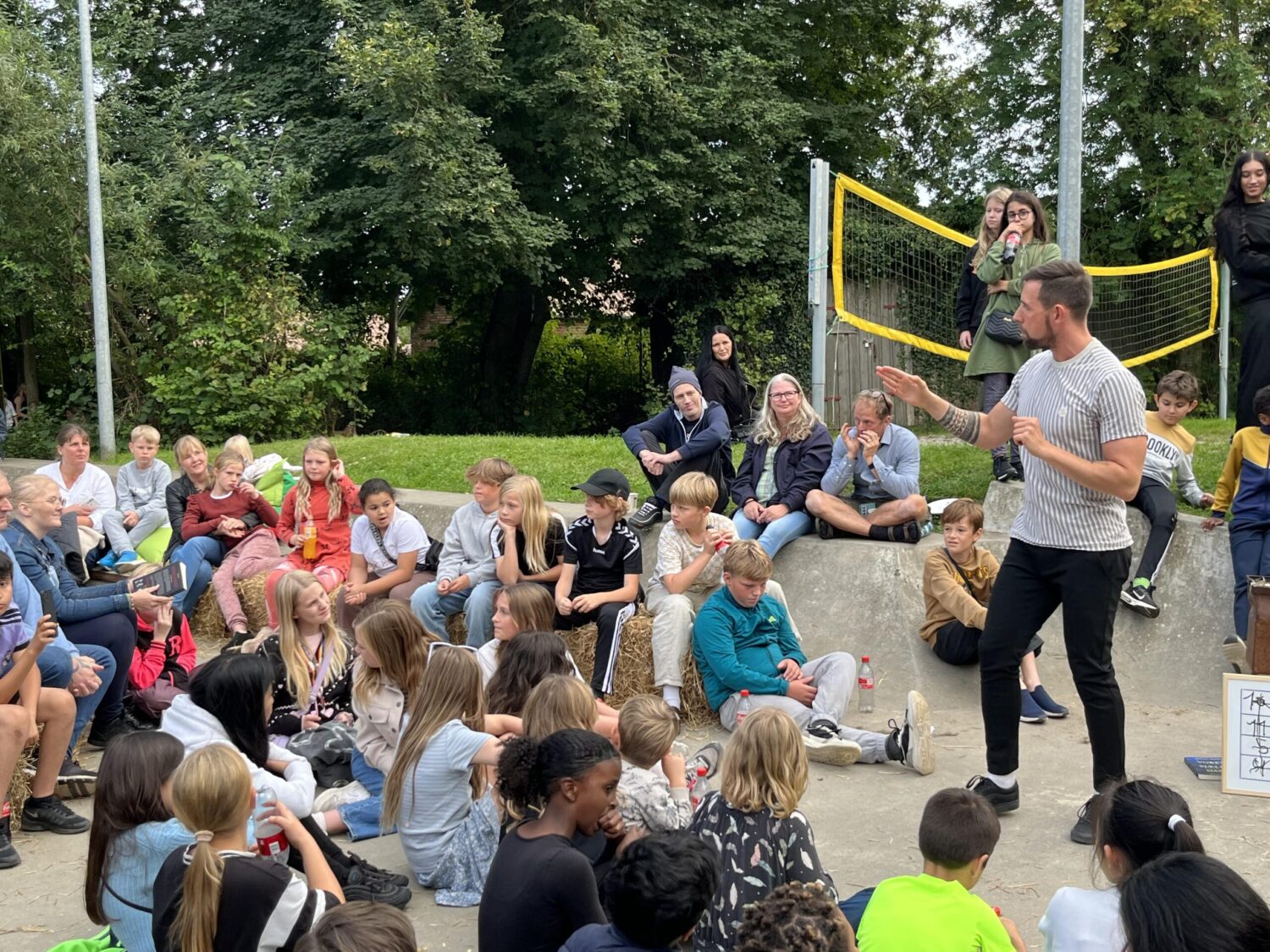 Mand står på en beachvolleybane og laver show foran en masse børn